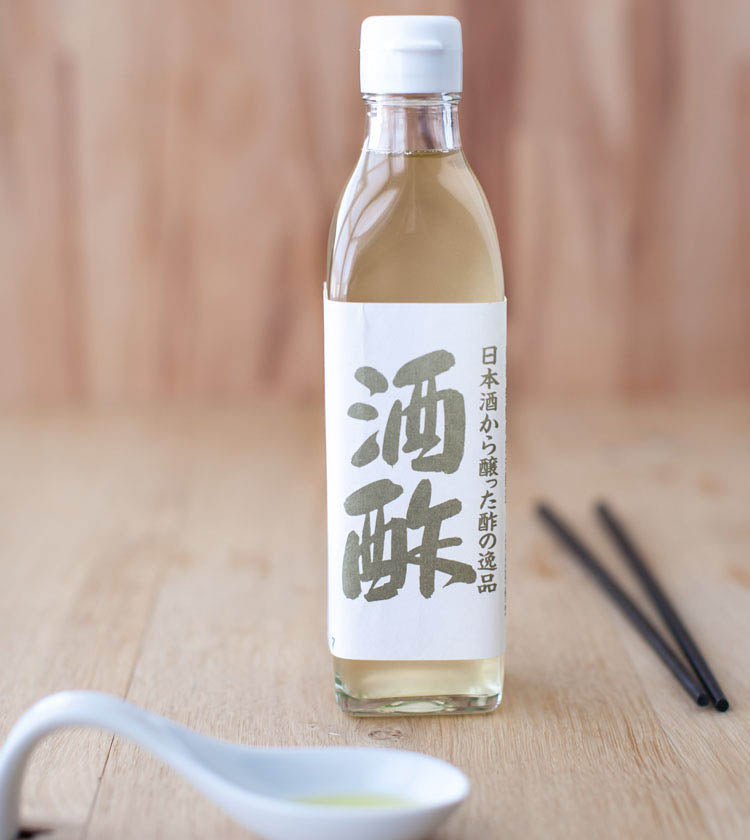 A clear glass bottle of Nigori Namazake Sakazu, Sake Vinegar with a white cap and Japanese label sits on a wooden surface beside black chopsticks and a white spoon.