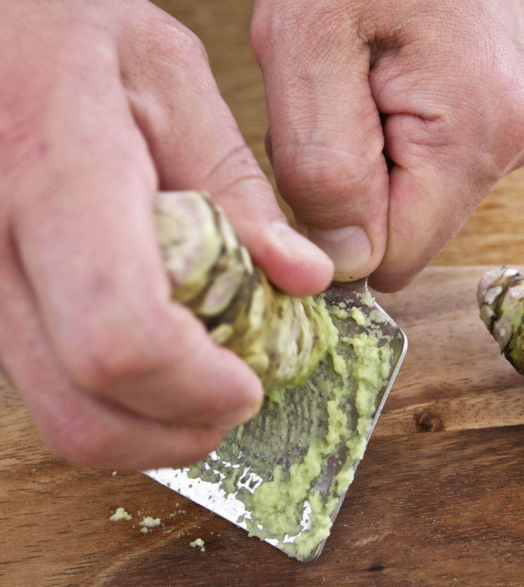Close-up of hands grating Small Fresh English Wasabi Rhizomes 100g on a metal grater over wood—ideal for adding authentic wasabi flavor to sushi.