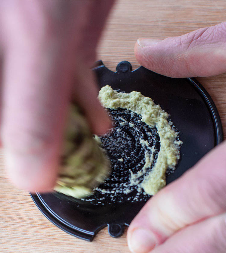 Using Small Fresh English Wasabi Rhizomes 200g, a person grinds the wasabi on a black grater atop wood, creating a naturally sweet paste.