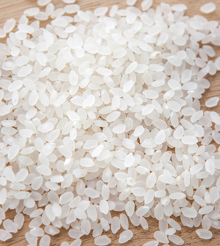 A pile of uncooked Akafuji Akitakomachi Rice grains is spread on a wooden surface, ideal for making sushi rice with a splash of Sushizu rice vinegar.