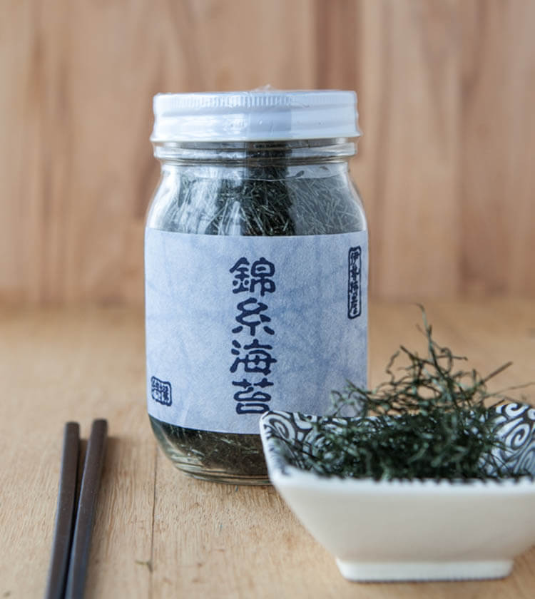 A glass jar of Nori Seaweed Angels Hair Cut - 30g with a Japanese label rests on a wooden table beside a dish of seaweed and chopsticks, showcasing this premium product from renowned Tôshi island.
