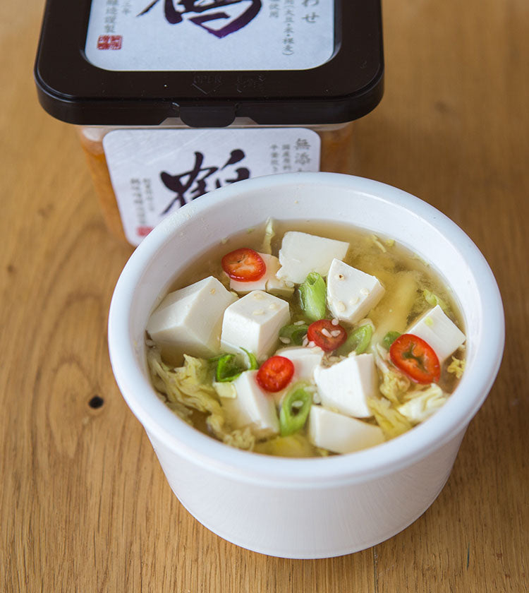 A bowl of soup with tofu, red chili, and green onion sits on wood, showcasing the umami richness of Awase Barley Miso - 500g featured in the background.