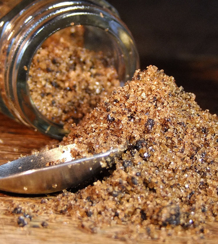 A jar of Black Garlic Sea Salt - 75g has tipped over, spilling its rich brown crystals onto a wooden surface beside a metal spoon, showcasing this unique seasoning’s distinct flavour.