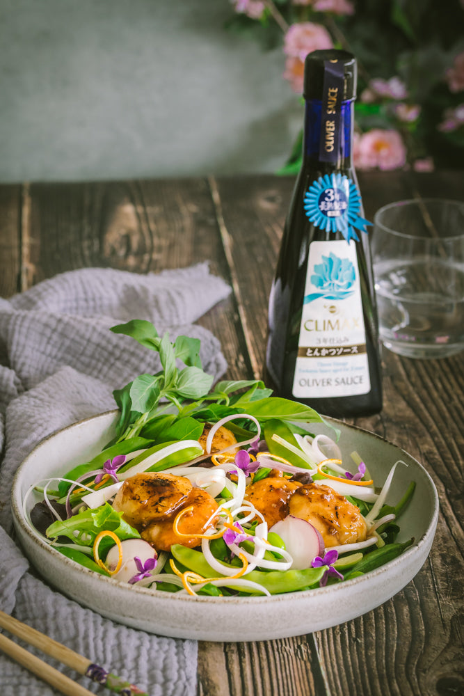A bowl of grilled chicken with vegetables and edible flowers sits beside a bottle of 3 Year Aged Tonkatsu Sauce - 350g on a wooden table with a napkin and chopsticks, evoking the flavors of Japanese street food.