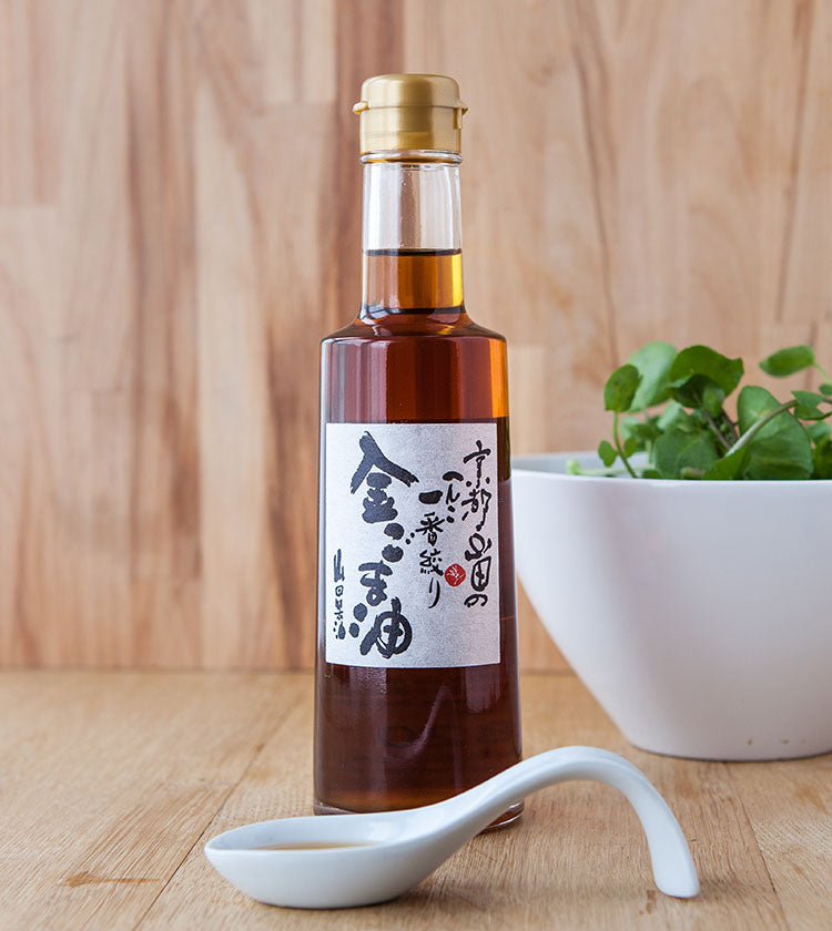 A 300ml glass bottle of Gold Sesame Oil with a gold cap sits on a wooden surface beside a white bowl of greens and a ceramic spoon, showcasing its rich aroma developed in wood-fired ovens.