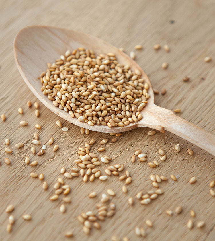 A wooden spoon filled with Wadaman Organic Roasted Gold Sesame Seeds rests on a wooden surface, with some seeds scattered around.