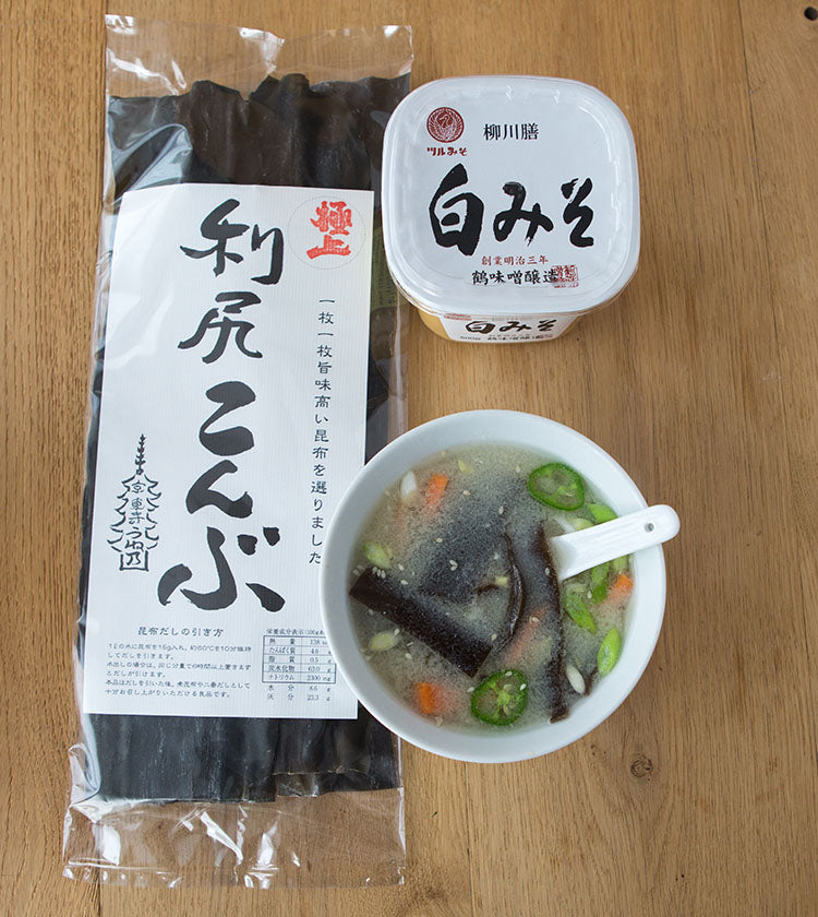 A package of Rishiri Wild Kombu Seaweed, a container of white miso paste, and a bowl of vegetable miso soup with kombu strips sit on a wooden surface.