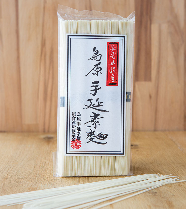 A 250g package of Thin Wheat Somen Noodles, featuring black and red kanji text, rests on a wooden surface with several loose noodles beside it.
