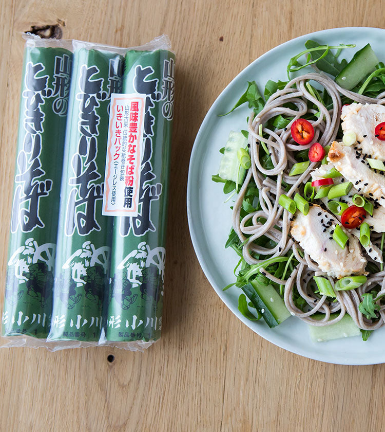 Three packs of Soba Noodles - 3 x 150g sit beside a plate of soba noodle salad topped with grilled chicken, red chili, cucumber, green onions, and greens.