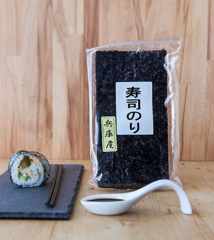 A package of Ariake Nori - 20 Half Sheets stands upright beside a sushi roll, chopsticks, and a spoonful of soy sauce on a wooden surface.