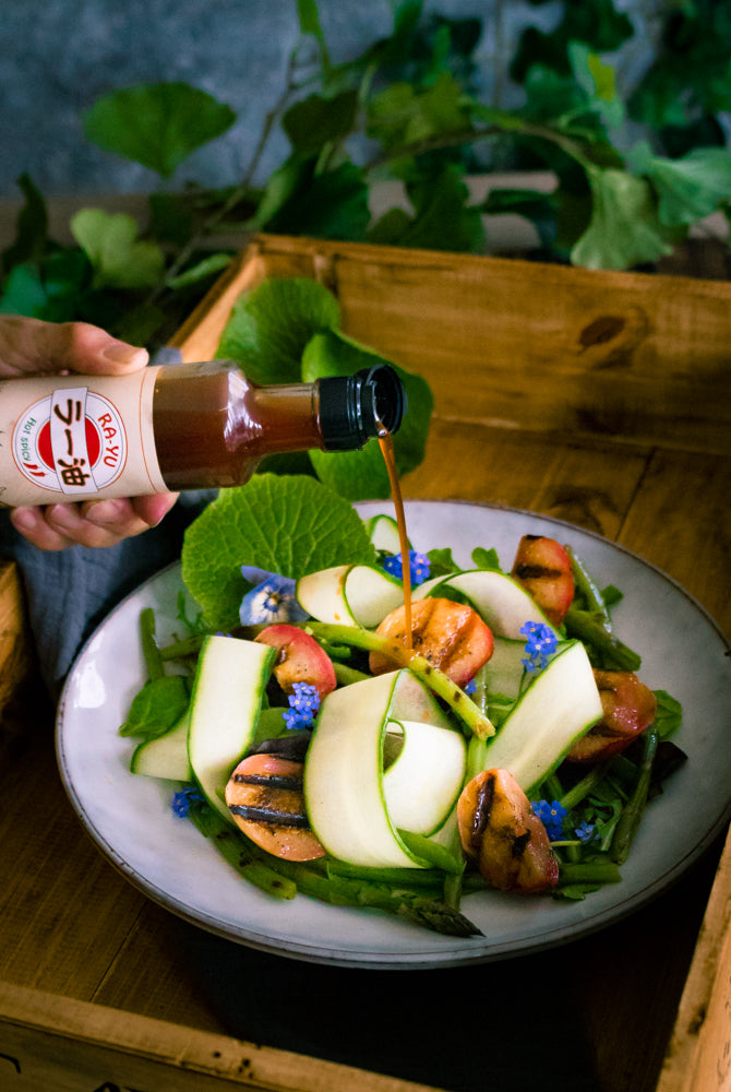 A hand pours Spicy Ra-Yu Pepper Sauce - 200g from a bottle onto a salad with grilled vegetables, fresh greens, zucchini ribbons, and small blue edible flowers.