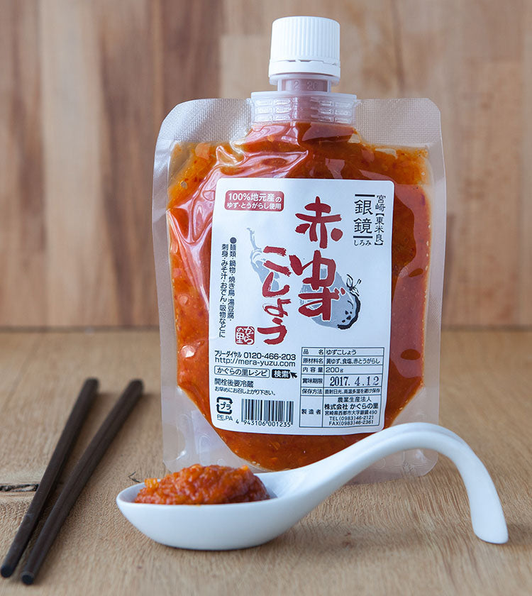 A pouch of Red Yuzu Kosho - 200g stands upright, with a white spoon holding the chili-infused condiment in front and a pair of chopsticks on the wooden surface.