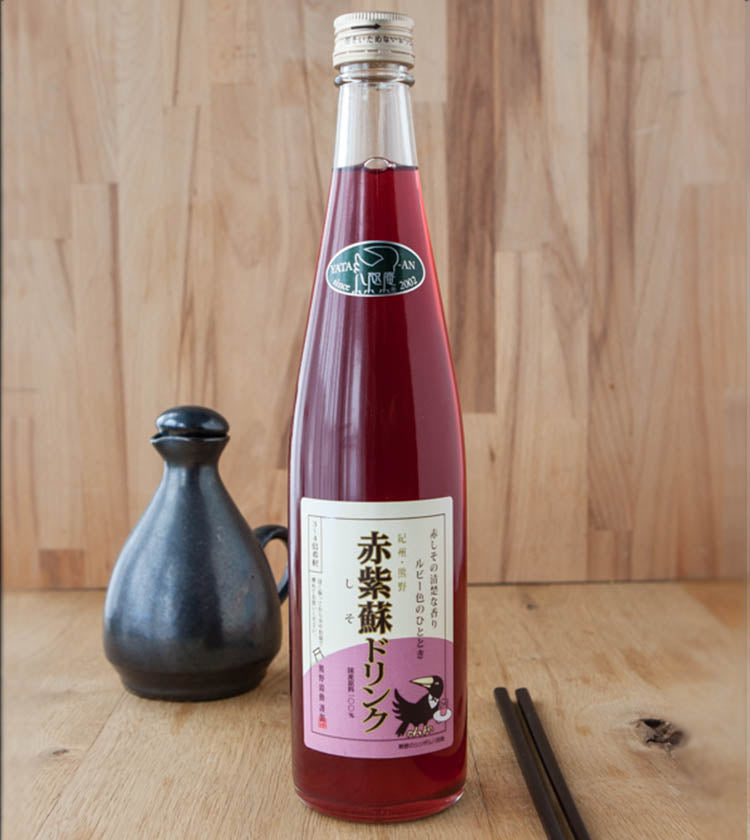 A bottle of Mikan Juice with Shiso - 500ml stands on a wooden surface, next to a black ceramic container and a pair of chopsticks.