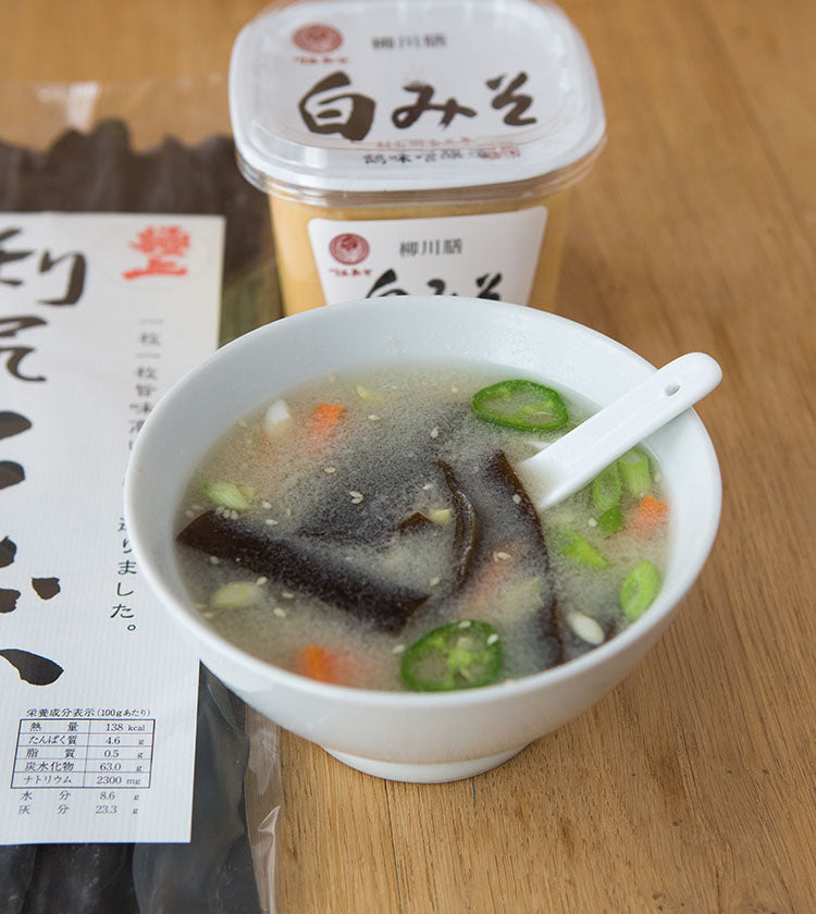 A bowl of miso soup with vegetables and seaweed, a white ceramic spoon, a container of White Miso - 500g, and a packet of dried seaweed arranged on a wooden surface showcasing rich umami flavour.