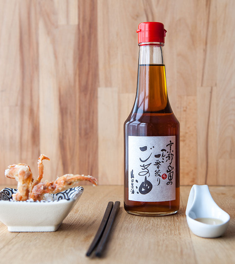 A 300ml bottle of White Sesame Oil with a label sits on a wooden surface beside fried food, chopsticks, and a small dish, evoking the rich flavors perfect for enhancing your favorite dishes.
