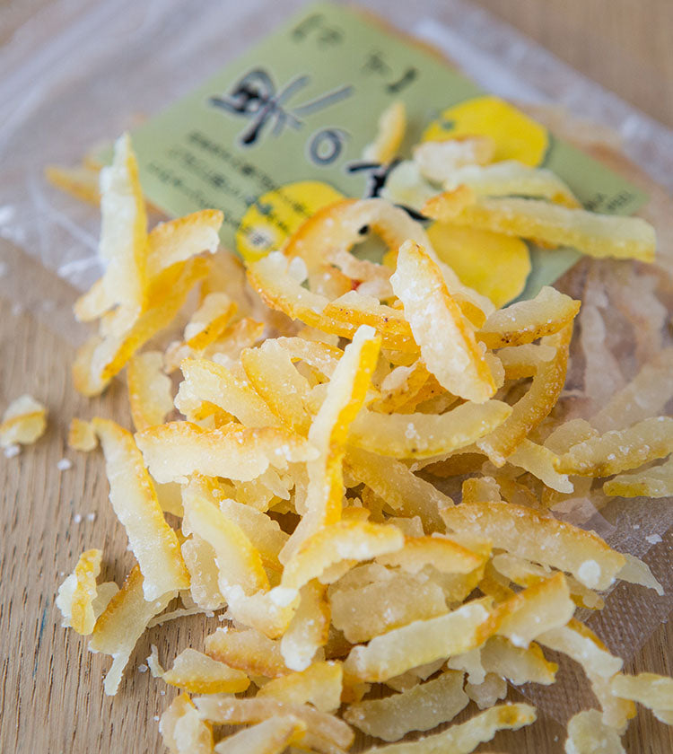 Candied Yuzu Peel, with a tart flavor and floral aroma, spills from a clear plastic package featuring a green and yellow label, shown on a wooden surface.