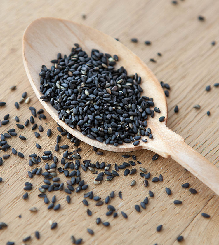 A wooden spoon filled with Wasabi Label Organic Black Sesame Seeds - 50g rests on a wooden surface, with extra black sesame seeds scattered nearby.