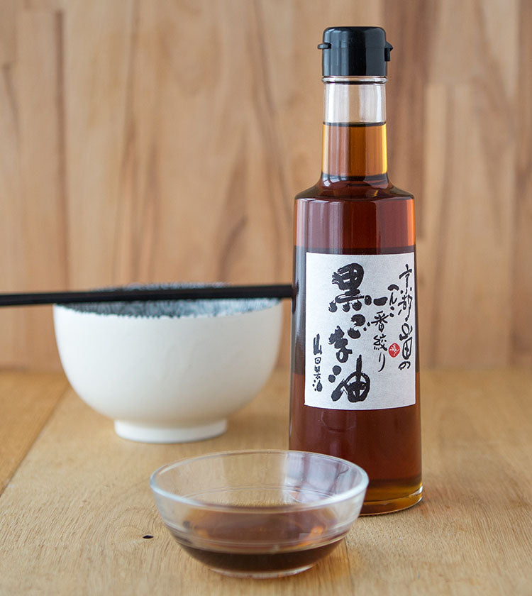 A 300ml bottle of Black Sesame Oil with a white label, a small glass dish of oil, and a white bowl with chopsticks are on a wooden surface.