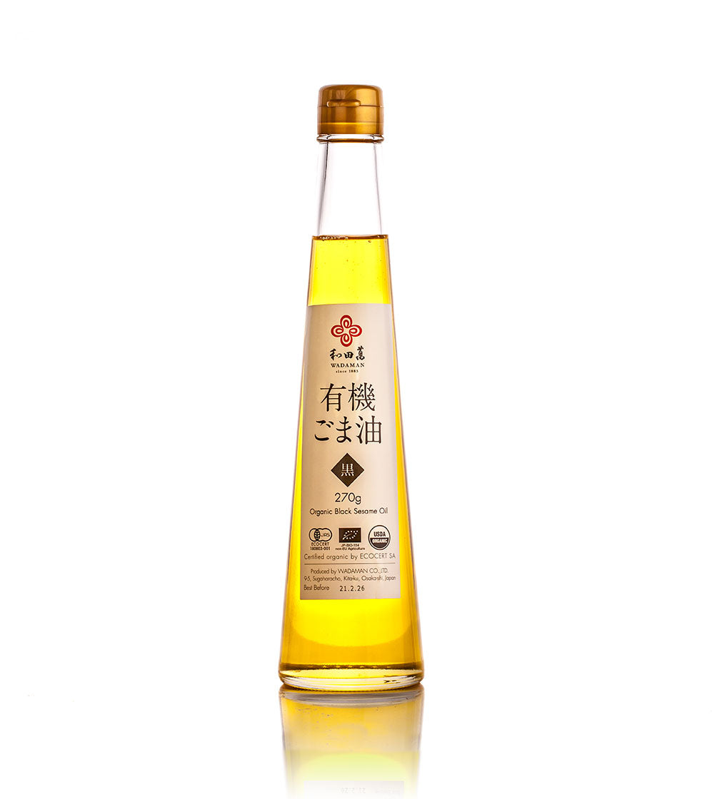 A clear glass bottle of Wadaman Organic Black Sesame Oil with a gold cap, Japanese labels, and organic roasted seeds stands upright on a white background.