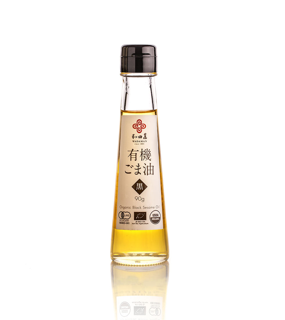 A clear glass bottle of Wadaman Organic Black Sesame Oil, made from roasted seeds, with a black cap and Japanese label, stands upright on a white background.
