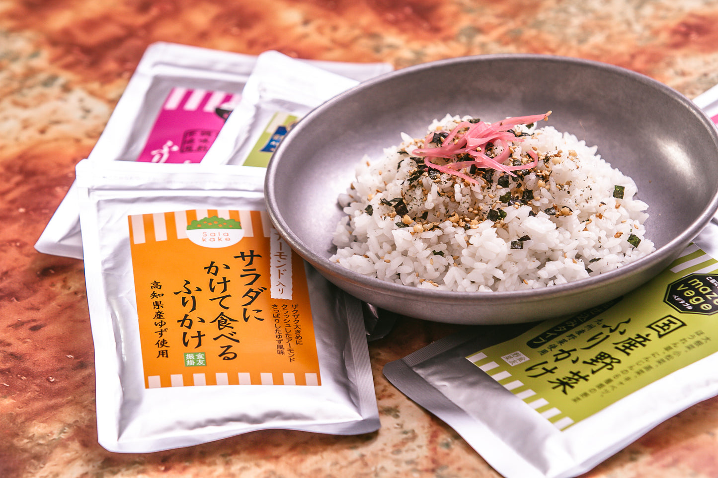 A bowl of white rice, topped with Furikake with Sesame, Ume Plum & Soy Sauce (50g) and pickled ginger, sits on a marble surface surrounded by assorted Japanese seasoning packets and a sprinkle of sesame seeds.