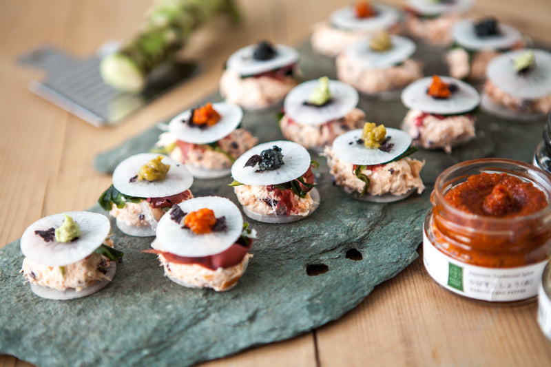 Several bite-sized canapés topped with white radish, yuzu peel, and colorful garnishes are arranged on a stone platter, with a jar of Black Yuzu Kosho - 50g and a grater visible in the background.