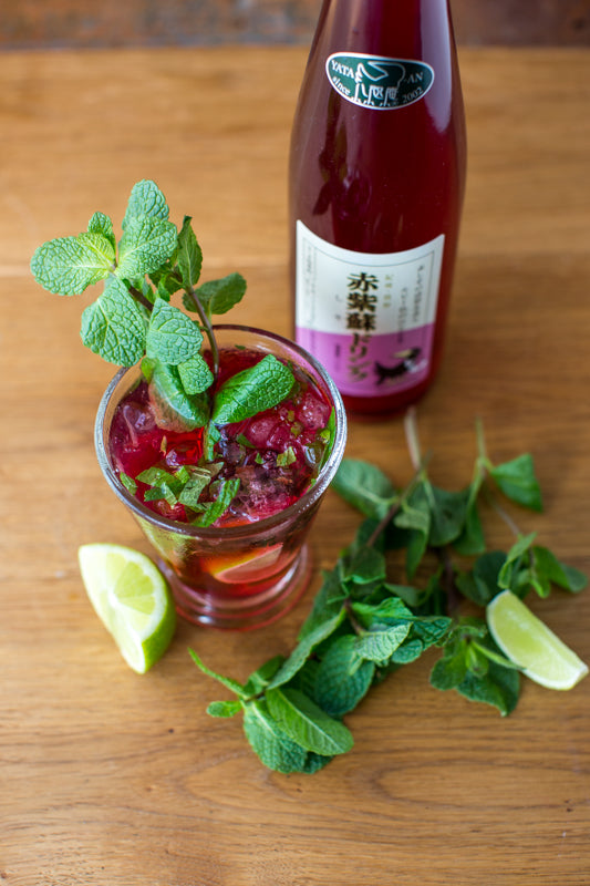 A glass of red cocktail garnished with lime wedges and mint leaves sits beside a 500ml bottle of Mikan Juice with Shiso on a wooden surface.