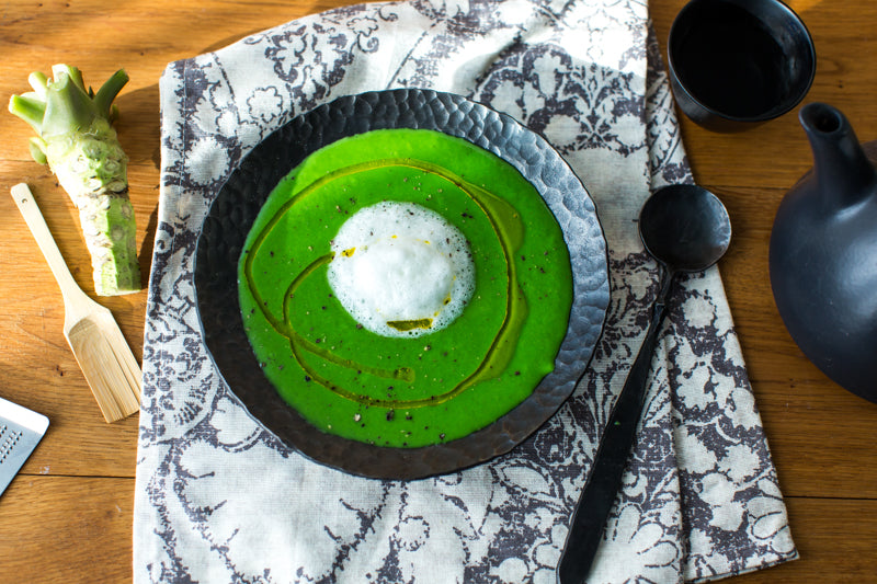 A black bowl of vibrant green soup with white foam and oil drizzle sits on patterned cloth, paired with a black spoon, teapot, cup, wasabi root, and enhanced by Nigori Namazake Sakazu Sake Vinegar for extra depth.