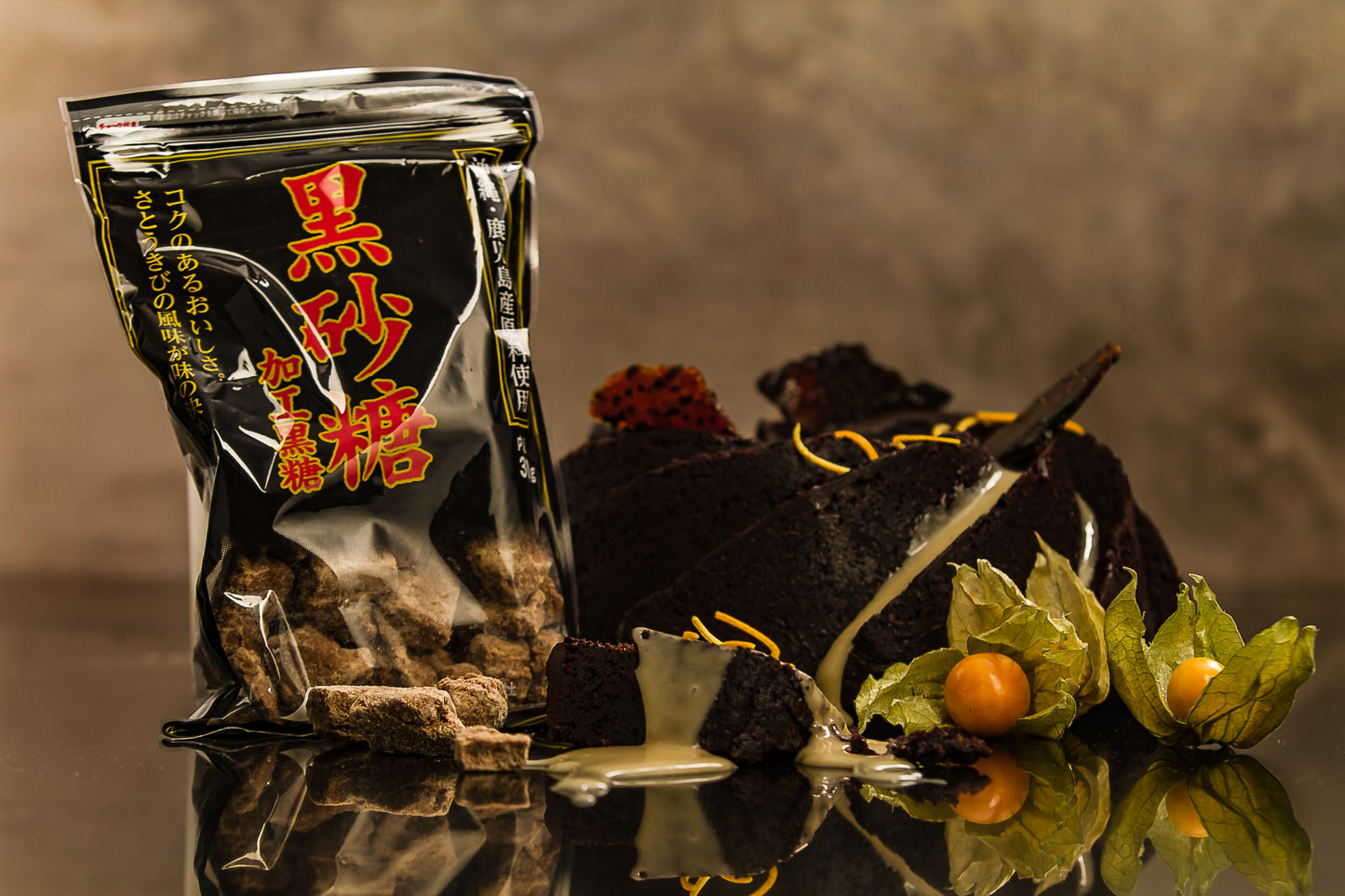 A 300g package of Brown Sugar from Okinawa & Kagoshima sits on a reflective surface alongside slices of chocolate cake with white icing and physalis fruits.