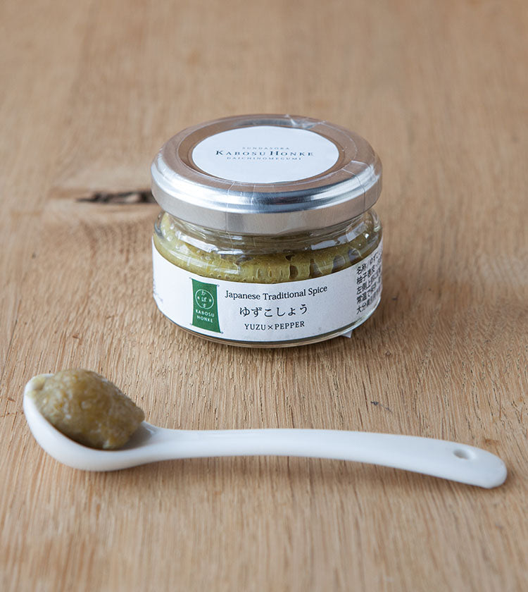 A small glass jar labeled Green Yuzu Kosho - 50g sits on a wooden surface. In front, a white ceramic spoon holds a dollop of the zesty green condiment, made from fresh chili and yuzu, popular in Japanese cuisine.