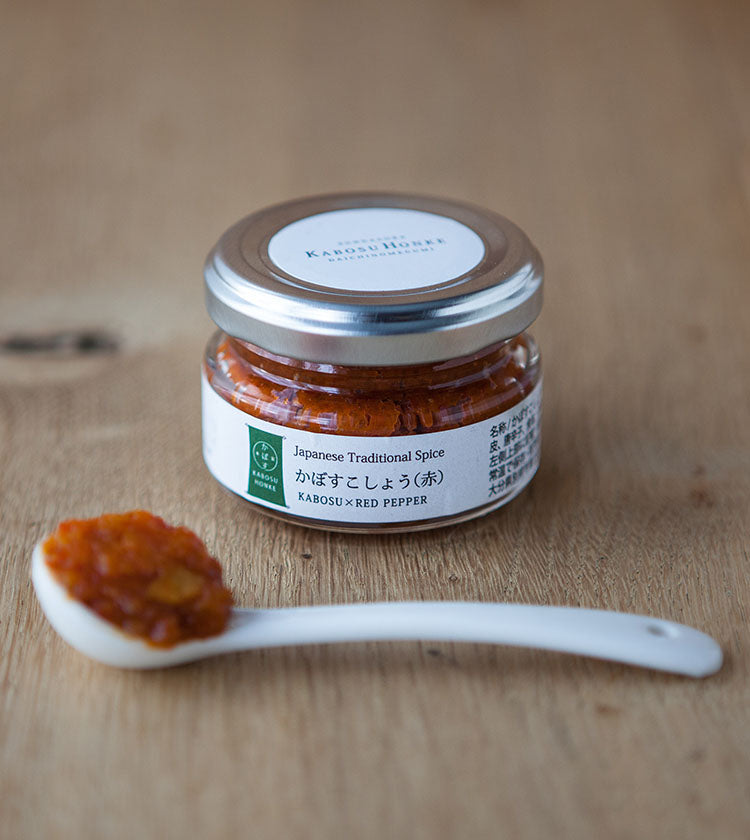 A small glass jar labeled “Red Kabosu Kosho - 50g On Offer” sits on a wooden surface, with a white spoon in front holding some of the zesty blend infused with Kabosu citrus and chillies.