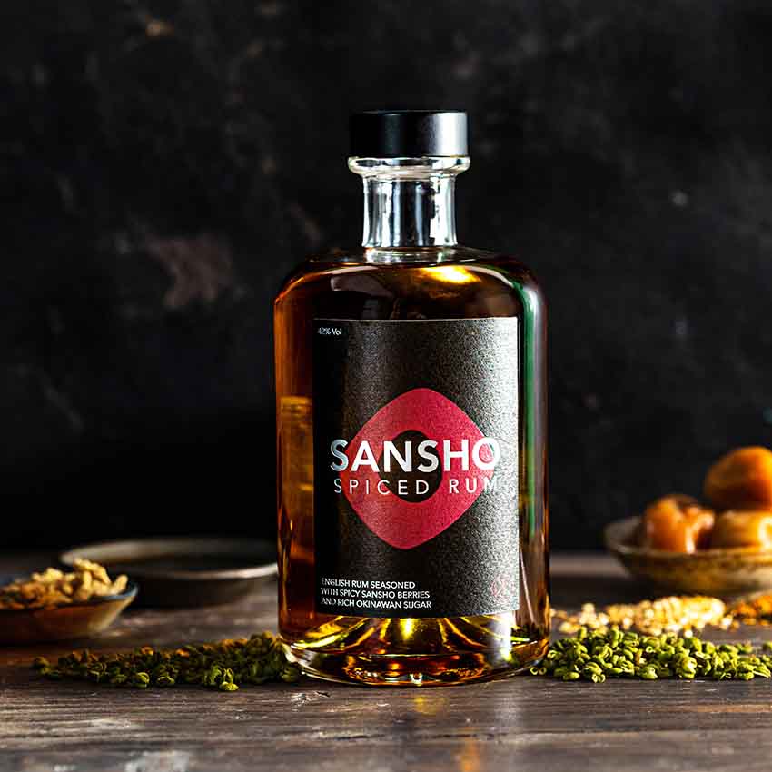 A bottle of Sansho Spiced Rum - 50cl sits on a dark surface, surrounded by spices, dried fruits, and Japanese-inspired Sansho Berries, set against a dramatic background.
