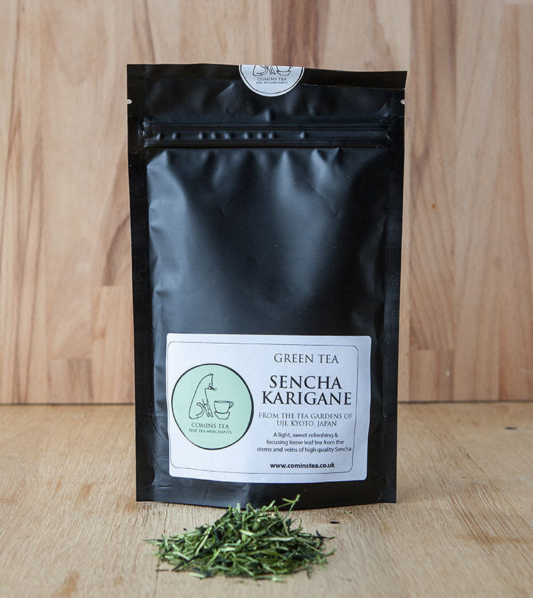 A black resealable bag labeled Sencha Karigane Tea - 50g stands on a wooden surface, with loose green tea leaves scattered in front.