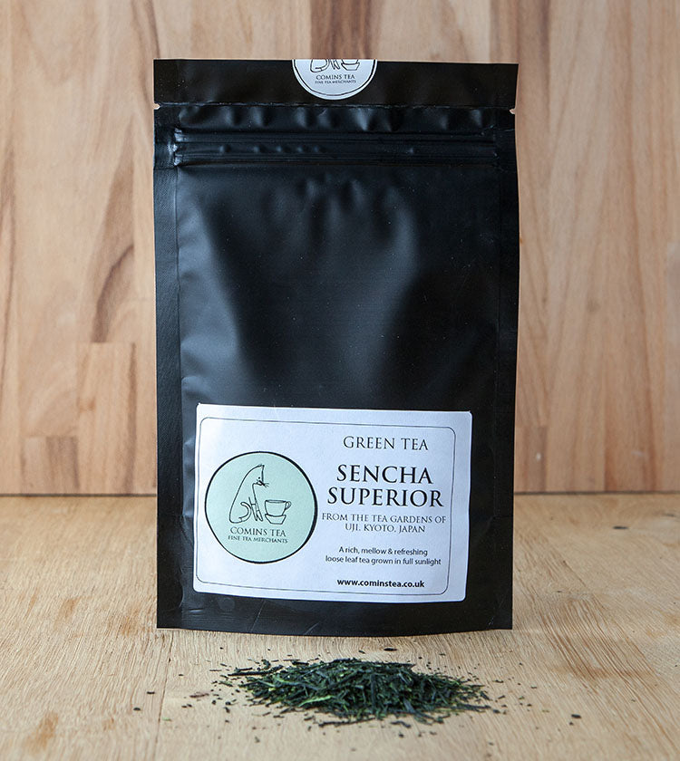 A black resealable bag of Sencha Superior Tea - 50g stands upright on a wooden surface, with loose tea leaves scattered in front.