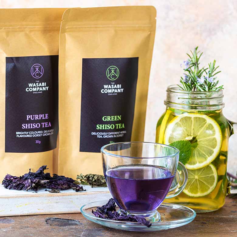 Two bags of Wasabi Company delicately flavoured tea, a glass cup with Purple Shiso Tea - 30g known for its calming effect, and a mason jar with lemon, herbs, and clear tea sit on the table.