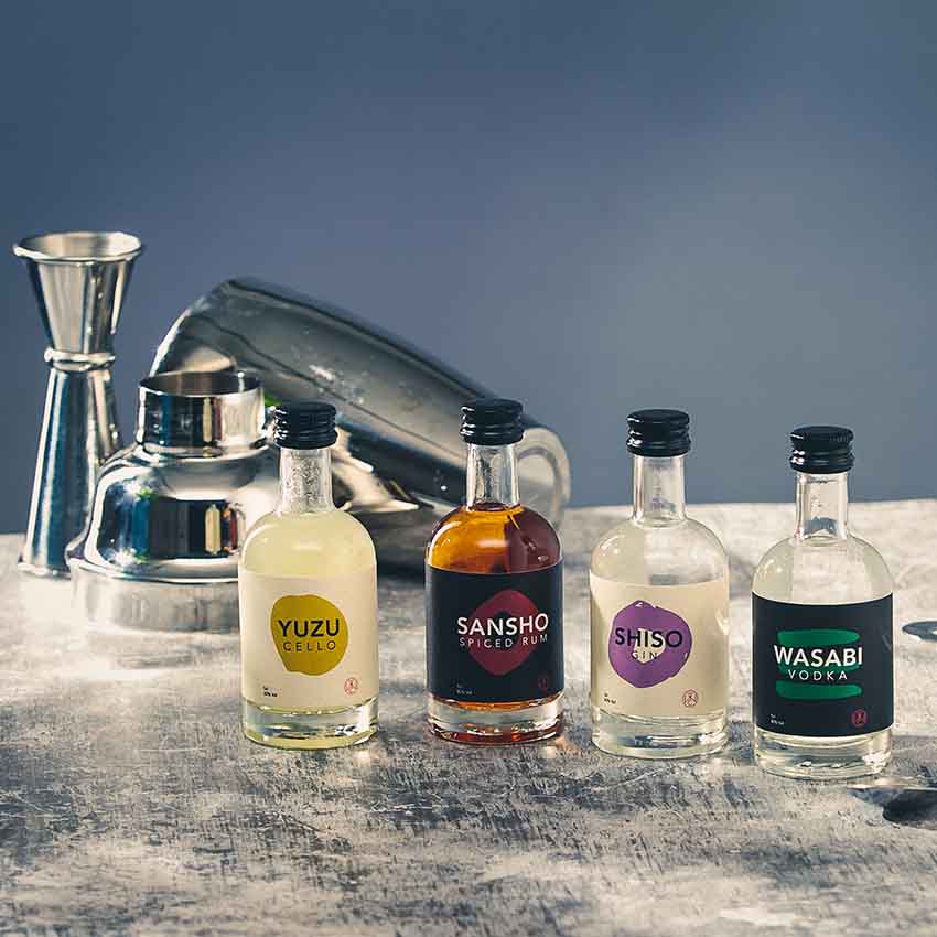 The Spirit of Japan – 4 x 5cl set features small bottles of Yuzu, Sansho, Shiso, and Wasabi Japanese spirits elegantly displayed with cocktail tools on a gray surface—an ideal gift for craft spirit lovers.