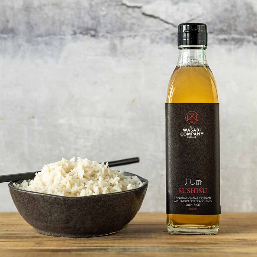 A bottle of Sushisu Sushi Rice Vinegar with Mirin (300ml) stands next to a bowl of cooked white rice with chopsticks, ready to add authentic flavor from the Kumano Mountain region.