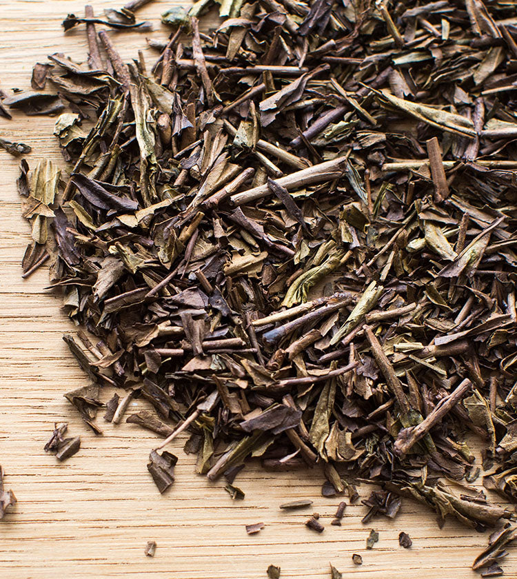 Houjicha Tea - 50g, featuring loose-leaf dried tea leaves pan roasted for a nutty flavour, is scattered on a light wooden surface.