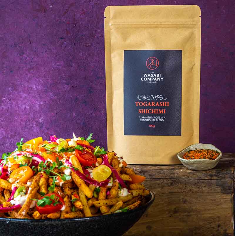 A bowl of seasoned fries with vegetables is placed before a Wasabi Label Togarashi Shichimi Spice Mix packet and a small dish of Japanese seven-flavour chilli blend, all set against a purple background.