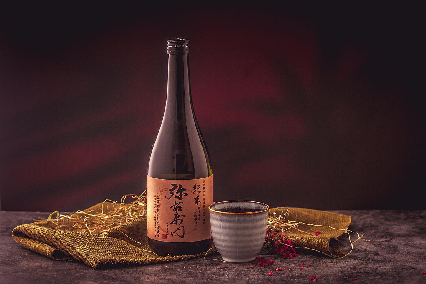 A bottle of Waning Moon Junmai Sake - 720ml with a Japanese label sits beside a filled ceramic cup on a fabric mat, accented by dried grass and set against a dark background—ideal for enjoying deep umami flavors.
