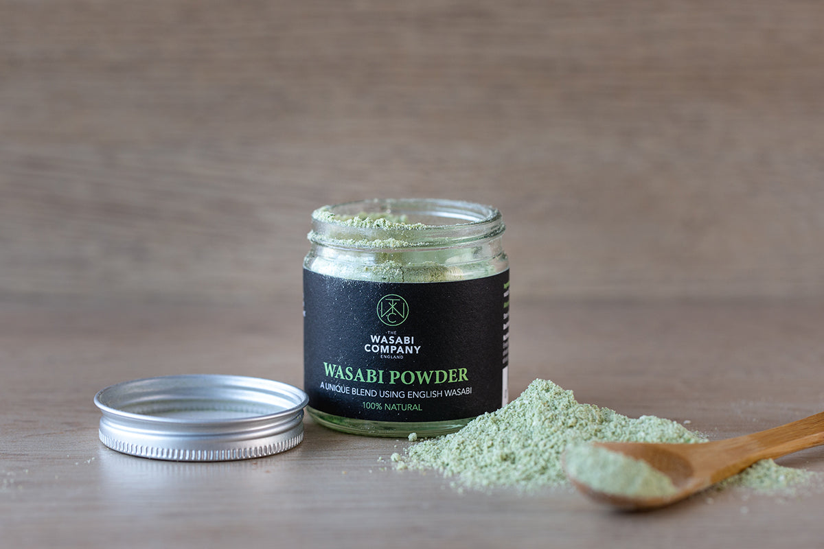 An open jar of Wasabi Powder - 23g with some powder spilled and a wooden spoon beside it. The lid sits next to the jar, ready to lock in the fresh Japanese spicy flavor.