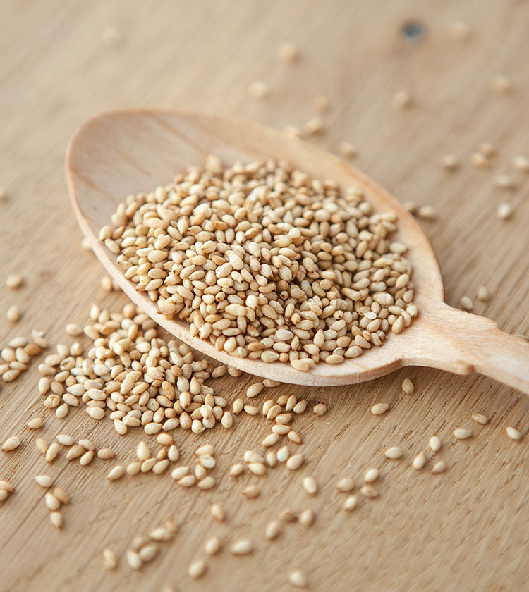 A wooden spoon holds Wadaman Organic Roasted White Sesame Seeds, with a few seeds scattered on a wooden surface.