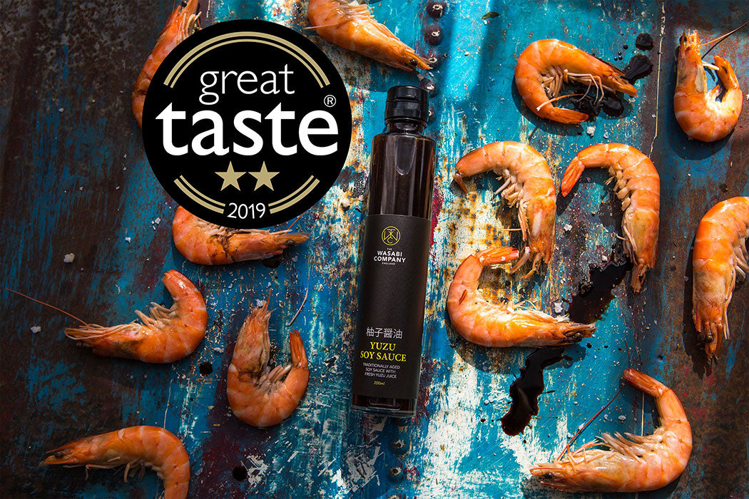 A 200ml bottle of Yuzu Soy Sauce, inspired by Japanese cuisine, is surrounded by cooked shrimp on a rustic blue surface, with a “Great Taste 2019” award badge displayed in the corner.