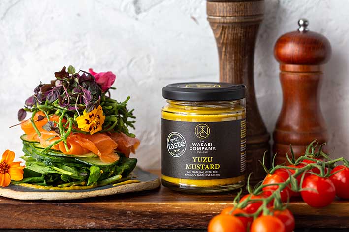 A 175g jar of award-winning Yuzu Mustard sits beside smoked salmon, greens, edible flowers, and cherry tomatoes—adding a burst of citrus flavor to every bite. A pepper grinder completes this elegant scene.