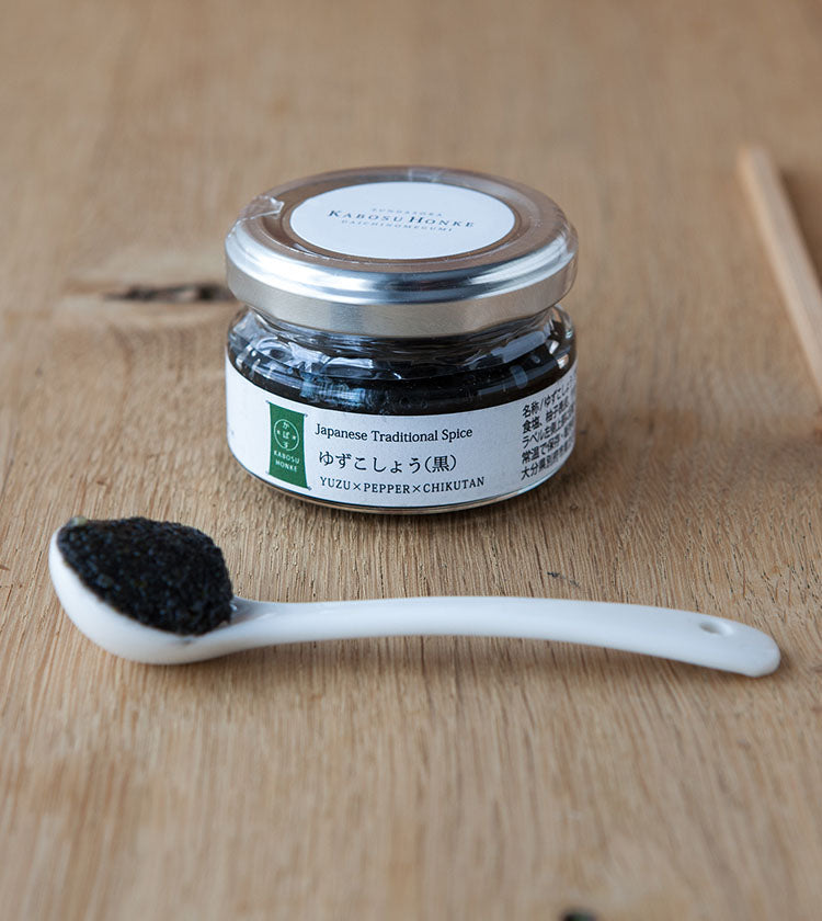 A 50g jar of Black Yuzu Kosho, a Japanese spice blend with yuzu peel, rests on a wooden surface beside a white spoon containing its dark paste.