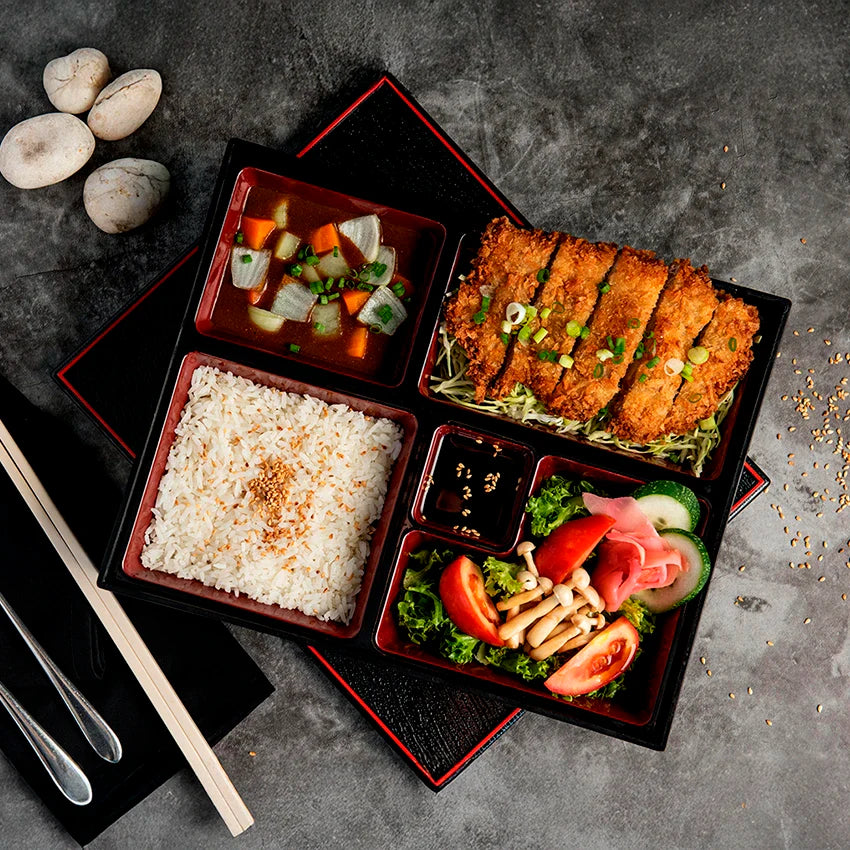 A bento box with white rice, miso soup, breaded cutlet, assorted vegetables, pickled ginger, dipping sauce, and garnishes on a gray surface with chopsticks and mushrooms nearby.