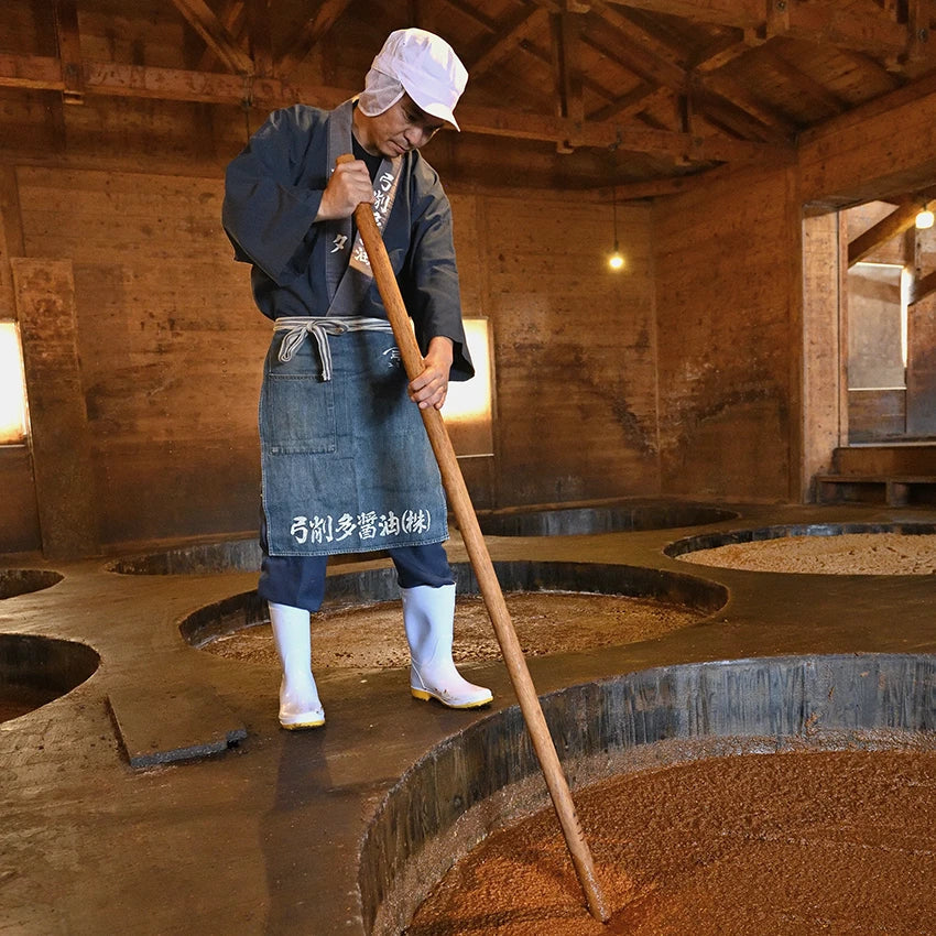A worker in traditional attire stirs a large vat of fermenting soy sauce in a wooden building.