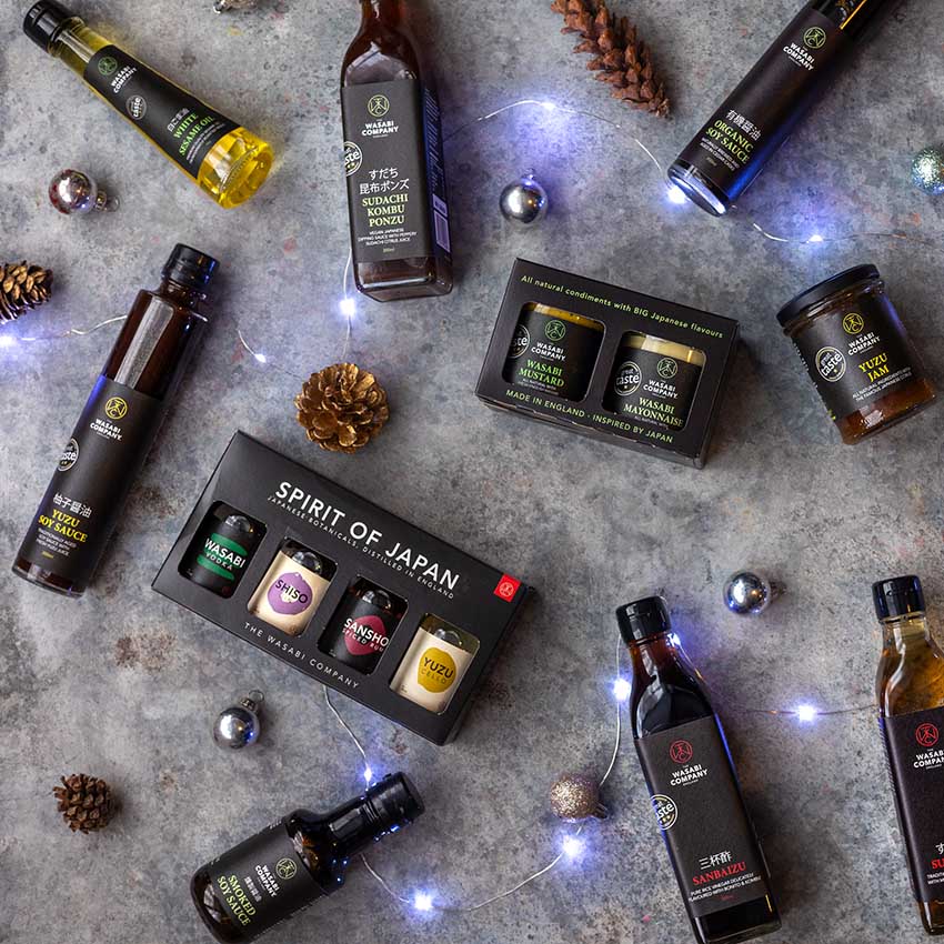 Assorted bottles and gift boxes of Japanese sauces and dressings are arranged on a gray surface with pinecones, ornaments, and string lights.