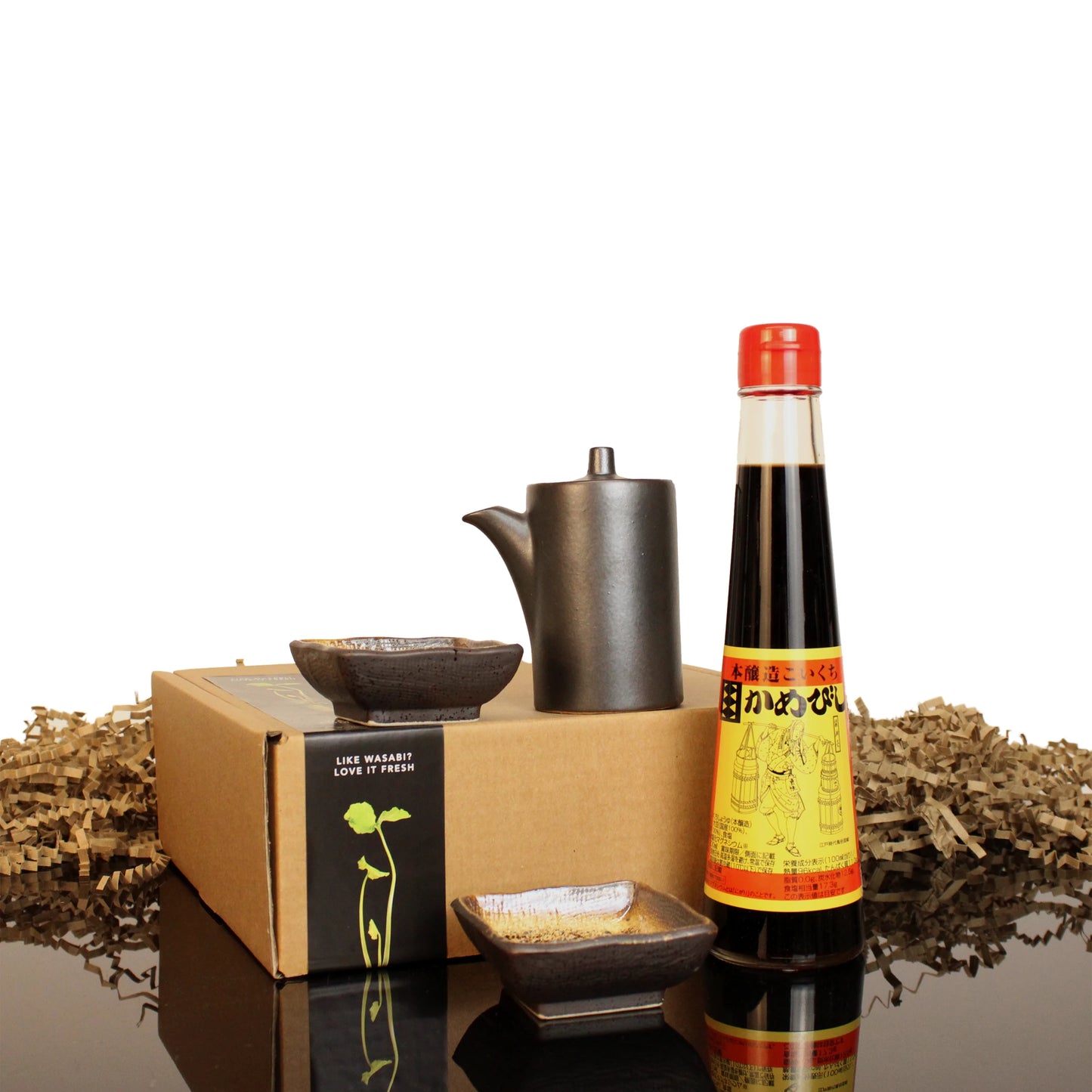 The 2 Year Aged Soy Sauce with Pourer & Serving Dishes is beautifully presented in a black ceramic pourer, two small dishes, and a box with shredded paper, making an elegant gift set displayed on a reflective surface.