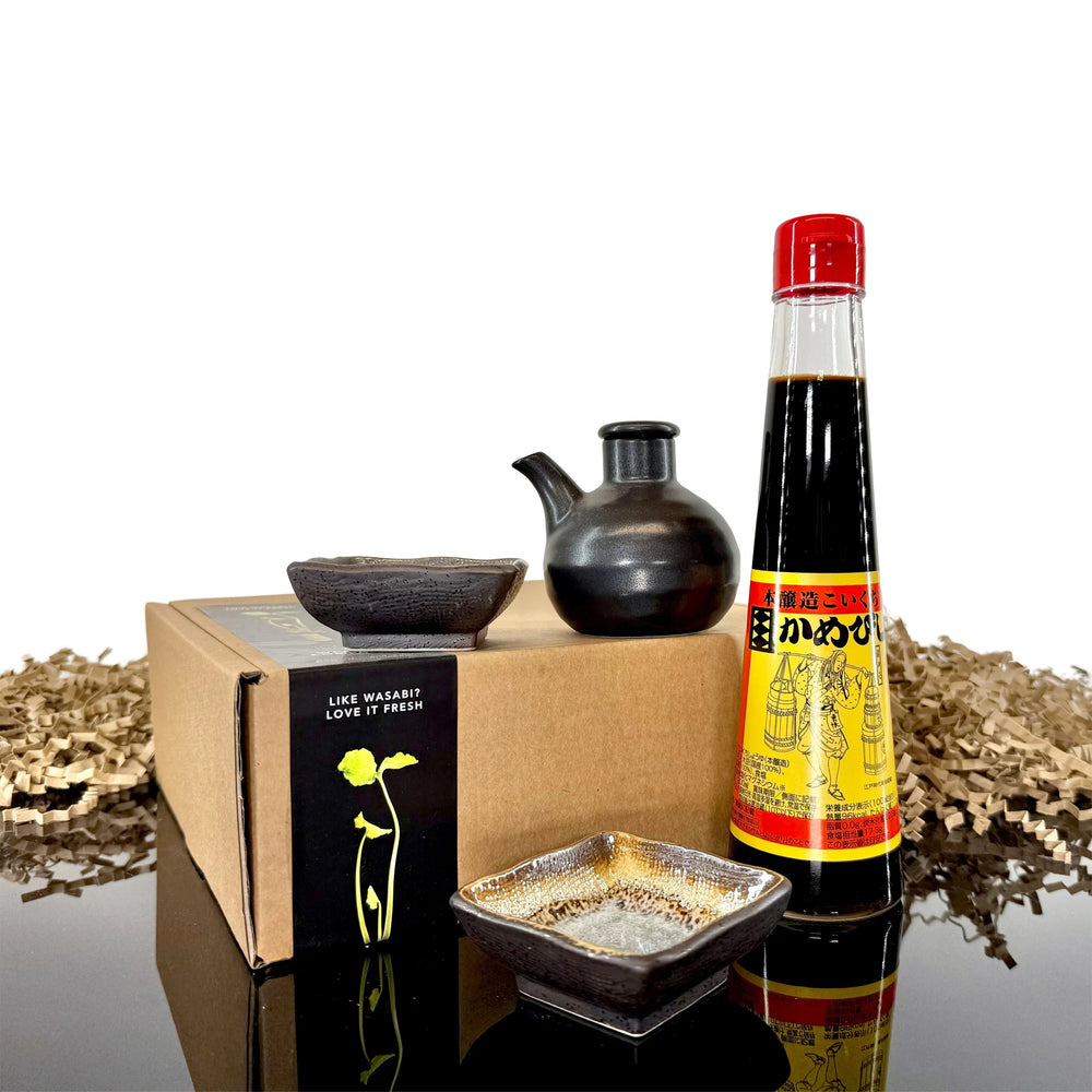 The 2 Year Aged Soy Sauce with Pourer & Serving Dishes is elegantly arranged on a black reflective surface, featuring the soy sauce bottle, ceramic pourer, two serving dishes, and wasabi-themed packaging.
